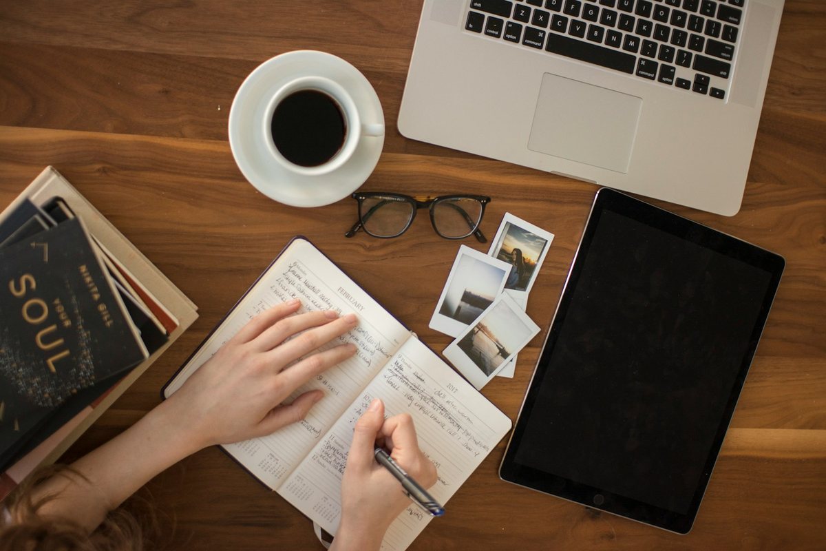Un bureau vu d’en haut : ordinateur, café, tablette et une main qui écrit dans un carnet