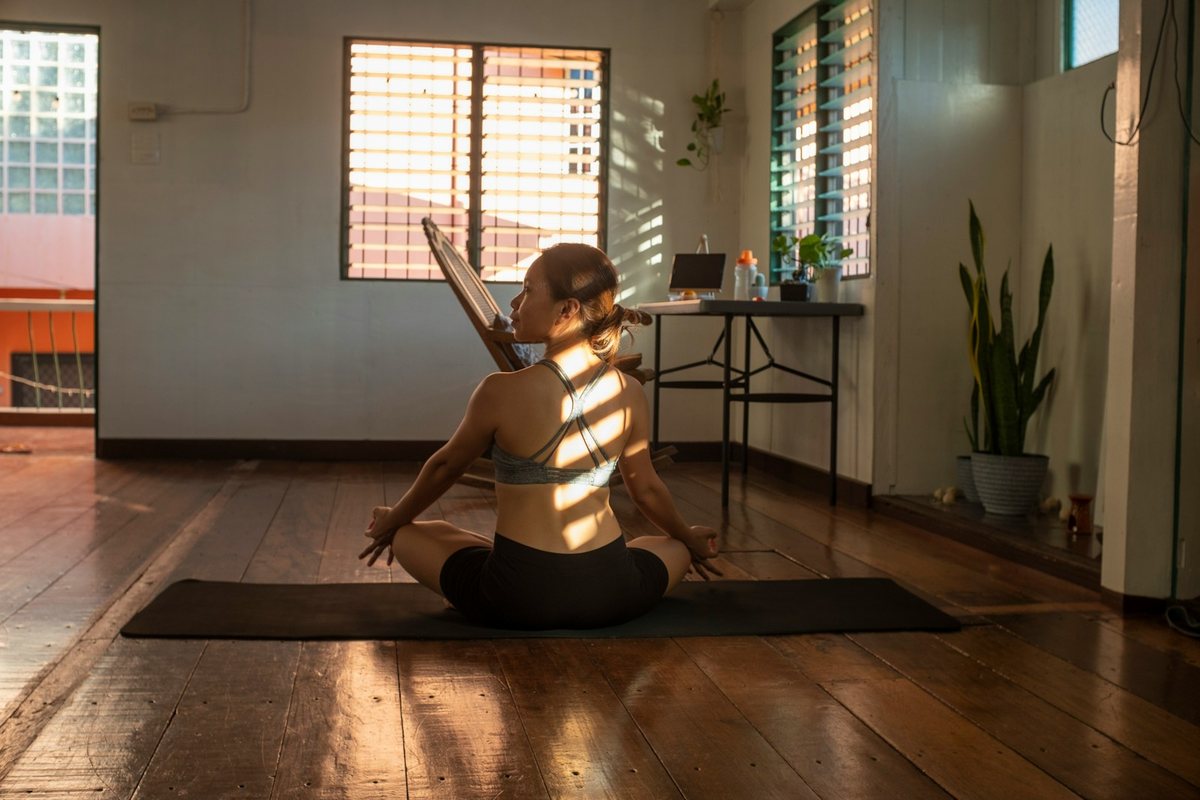 Une femme assise en tailleur sur un tapis de yoga, vue de dos, dans la lumière du matin